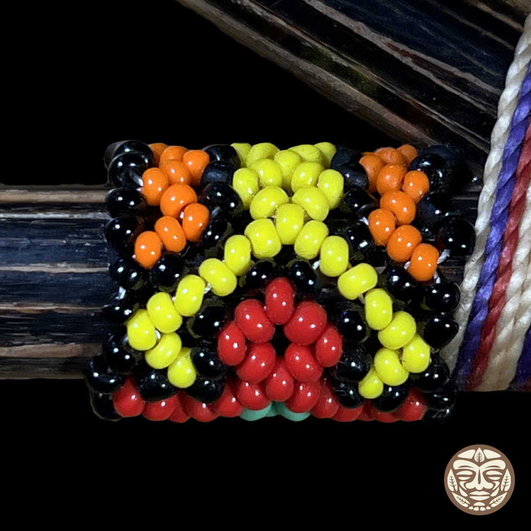 A close-up of a handcrafted beaded kuripe featuring a geometric pattern of black, orange, yellow, and red beads, shown wrapped around a dark palo chonta wood.