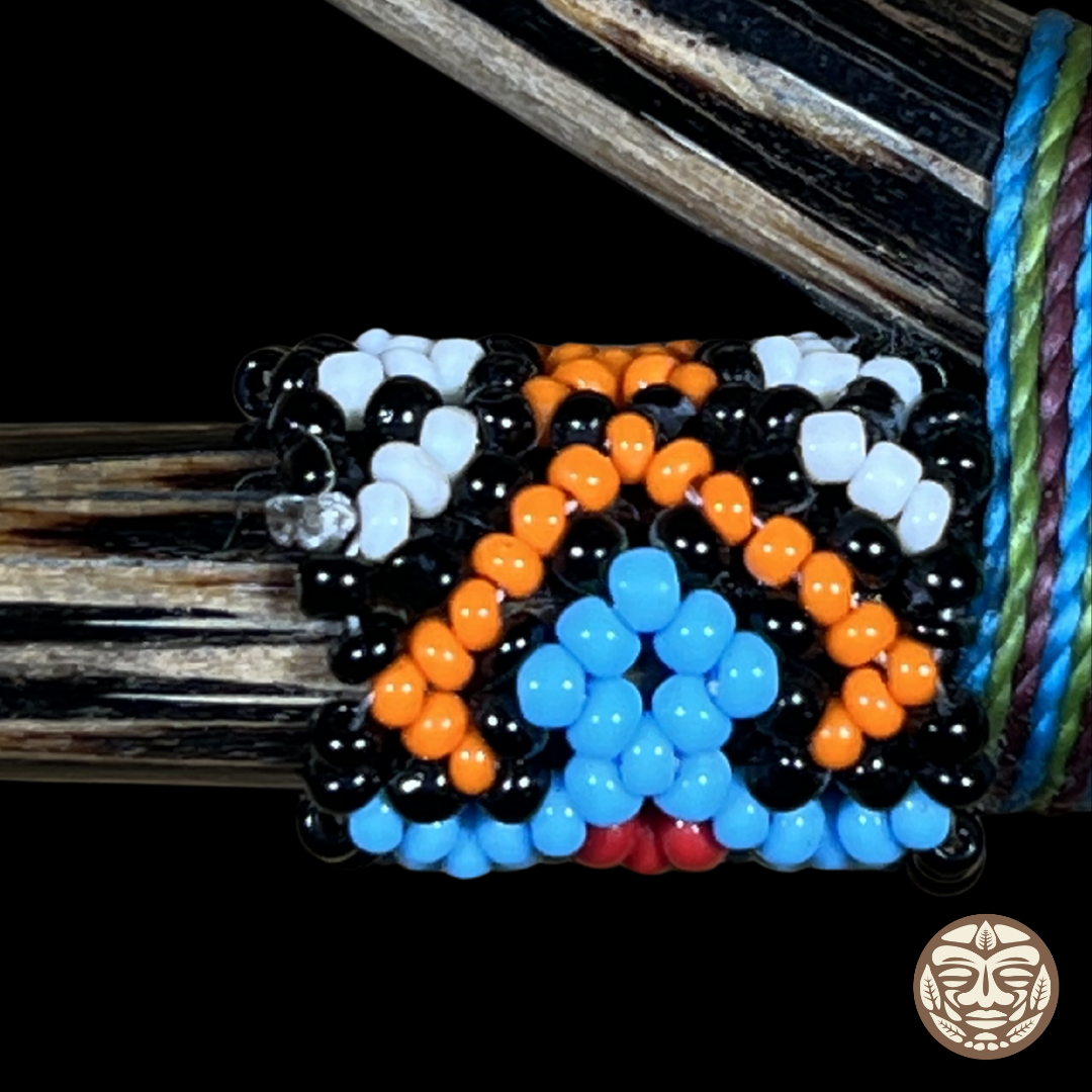 A close-up of a handcrafted beaded wrap kuripe attached to a palo chonta wood, featuring rows of glossy black, white, orange, and light blue beads in a geometric pattern. 