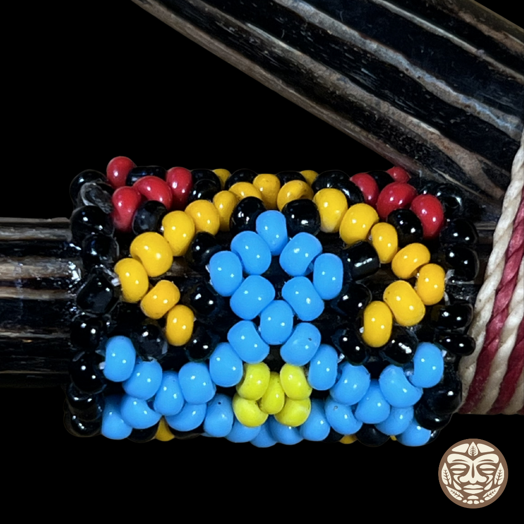 A close-up of a beaded wrap kuripe featuring a geometric pattern of black, yellow, blue, and red beads, wrapped around a dark palo chonta wood with a red-and-white cord on the right side.