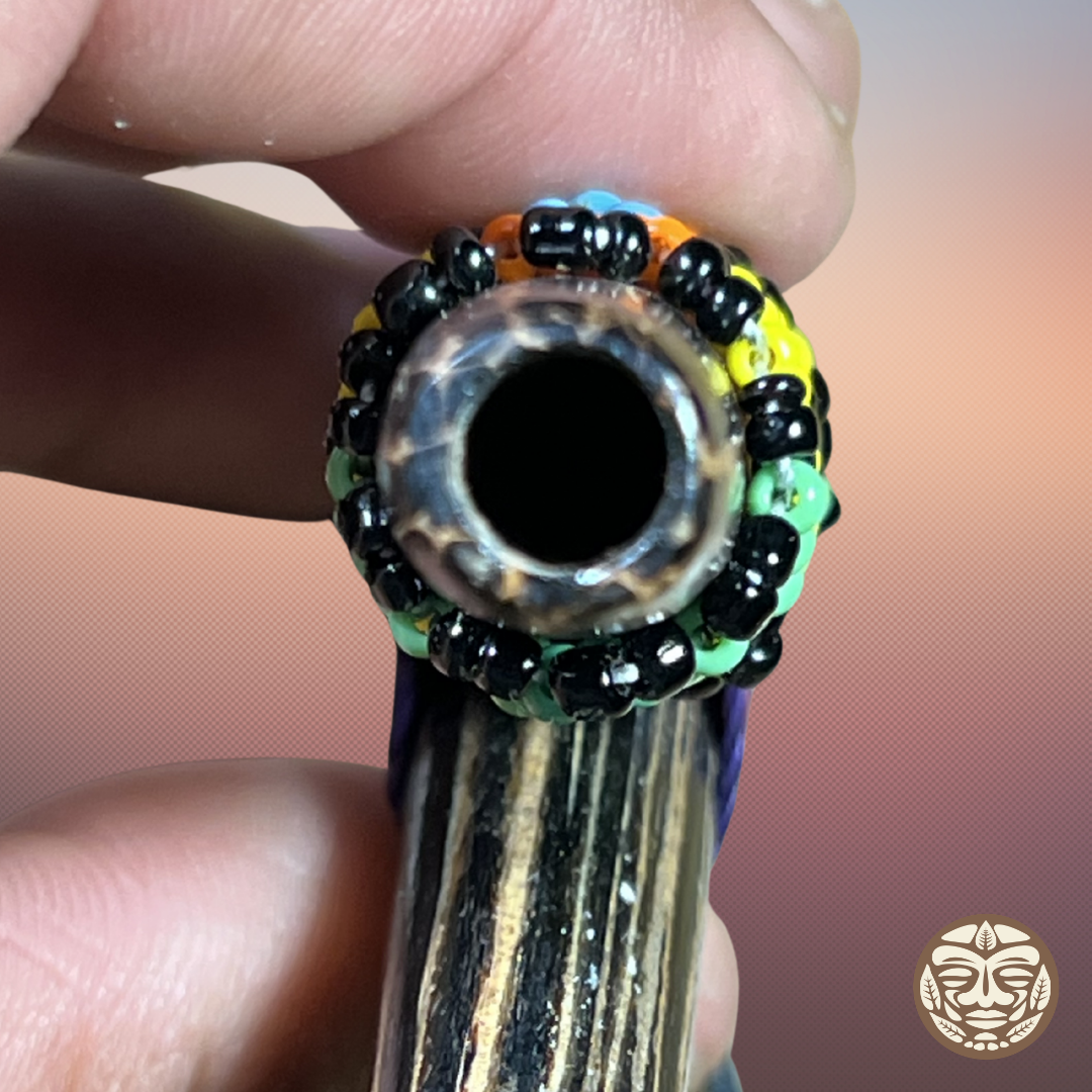 A close-up of a hand holding a carved palo chonta kuripe blow hole with a decorative beaded rim in black, green, yellow, orange, and blue.