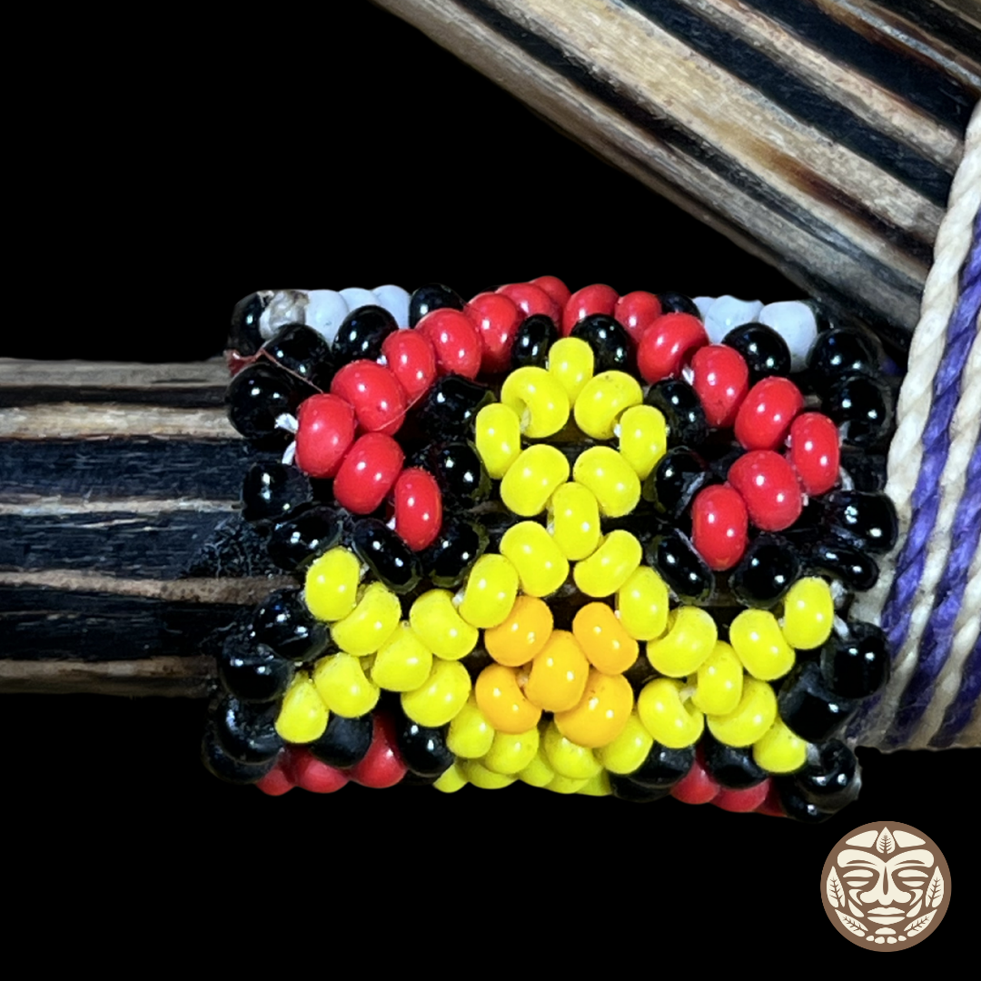A beaded wrap kuripe featuring a central yellow and orange sun-like motif surrounded by red, black, and white beads, shown wrapped around a dark palo chonta wood.  