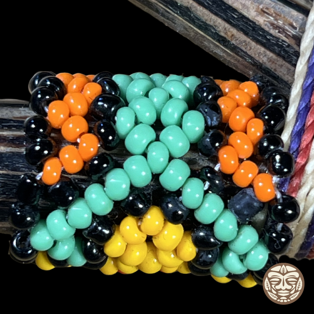 A close-up of a woven beaded kuripe with rows of glossy oval beads in mint green, bright orange, yellow, and black, arranged in a geometric pattern.